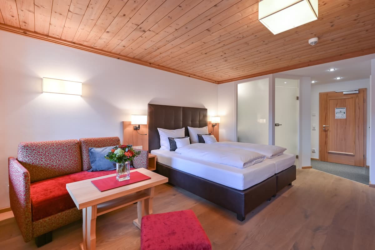 A modern hotel room with a wooden ceiling, a large bed, a sofa, a small table with a vase of flowers, and a nightstand.