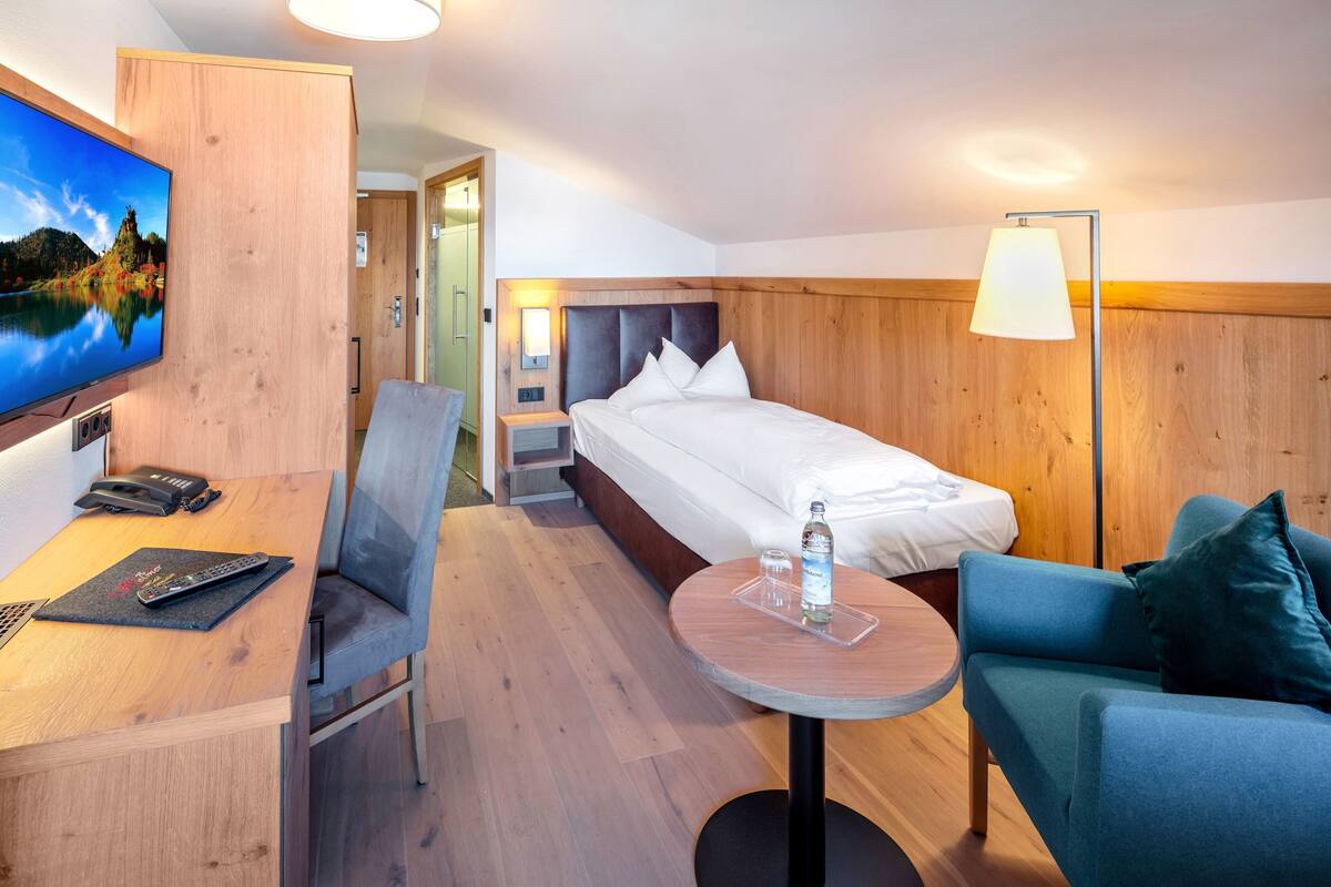 A hotel room with a bed, a desk with a chair, a TV, a lamp, and a small table with a bottle and glasses.