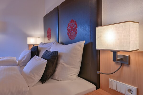 Una cama bien hecha con almohadas blancas y oscuras, un cabecero con motivos decorativos rojos, una lámpara de noche y un enchufe en la pared.