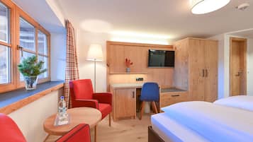A compact hotel room with a bed, a desk with a chair, a small table with a chair, a TV mounted on the wall, and a window with curtains.