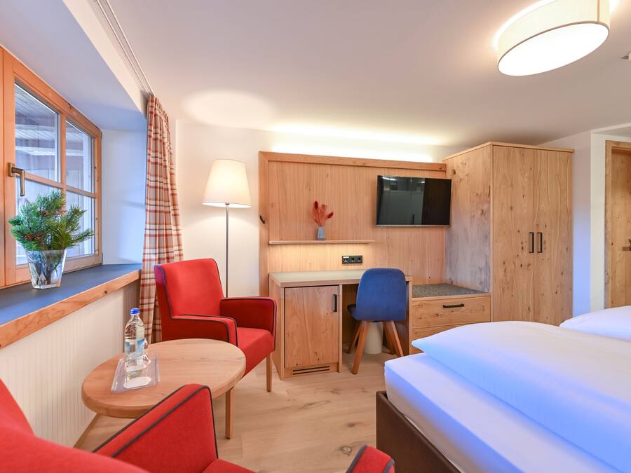 A compact hotel room with a bed, a desk with a chair, a small table with a chair, a TV mounted on the wall, and a window with curtains.