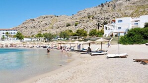 Una playa con tumbonas y sombrillas, una colina rocosa al fondo y edificios con balcones.