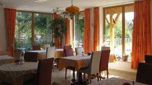 Un'area pranzo con tavoli e sedie, ampie finestre con tende arancioni e un lampadario a forma di fiori.