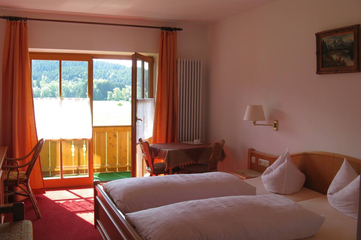 Une chambre d’hôtel avec deux lits, une table à manger, des chaises, une fenêtre avec des rideaux, un radiateur et une lampe fixée au mur.