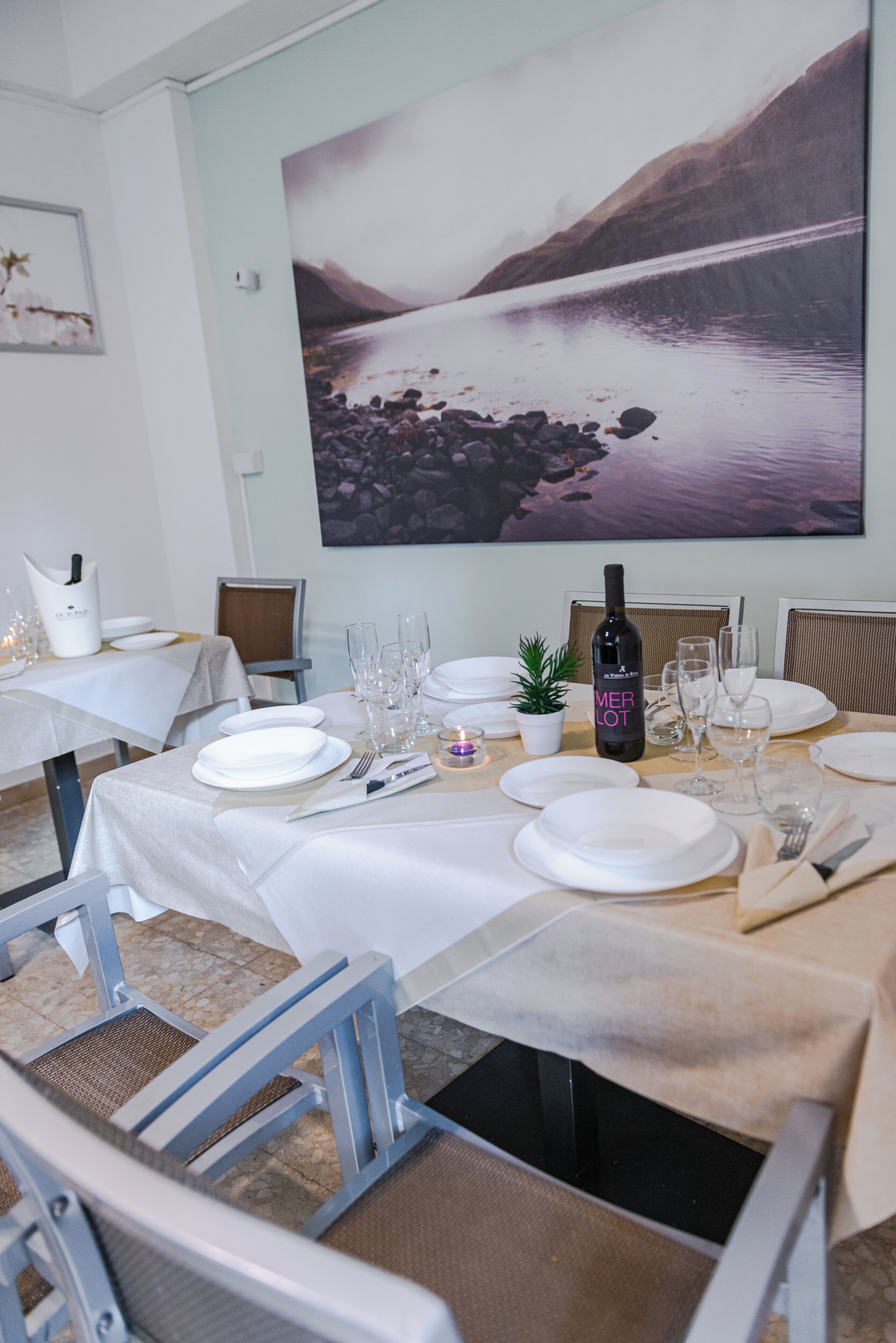 A dining area with a table set for six, a bottle of Merlot, and a large scenic painting on the wall.