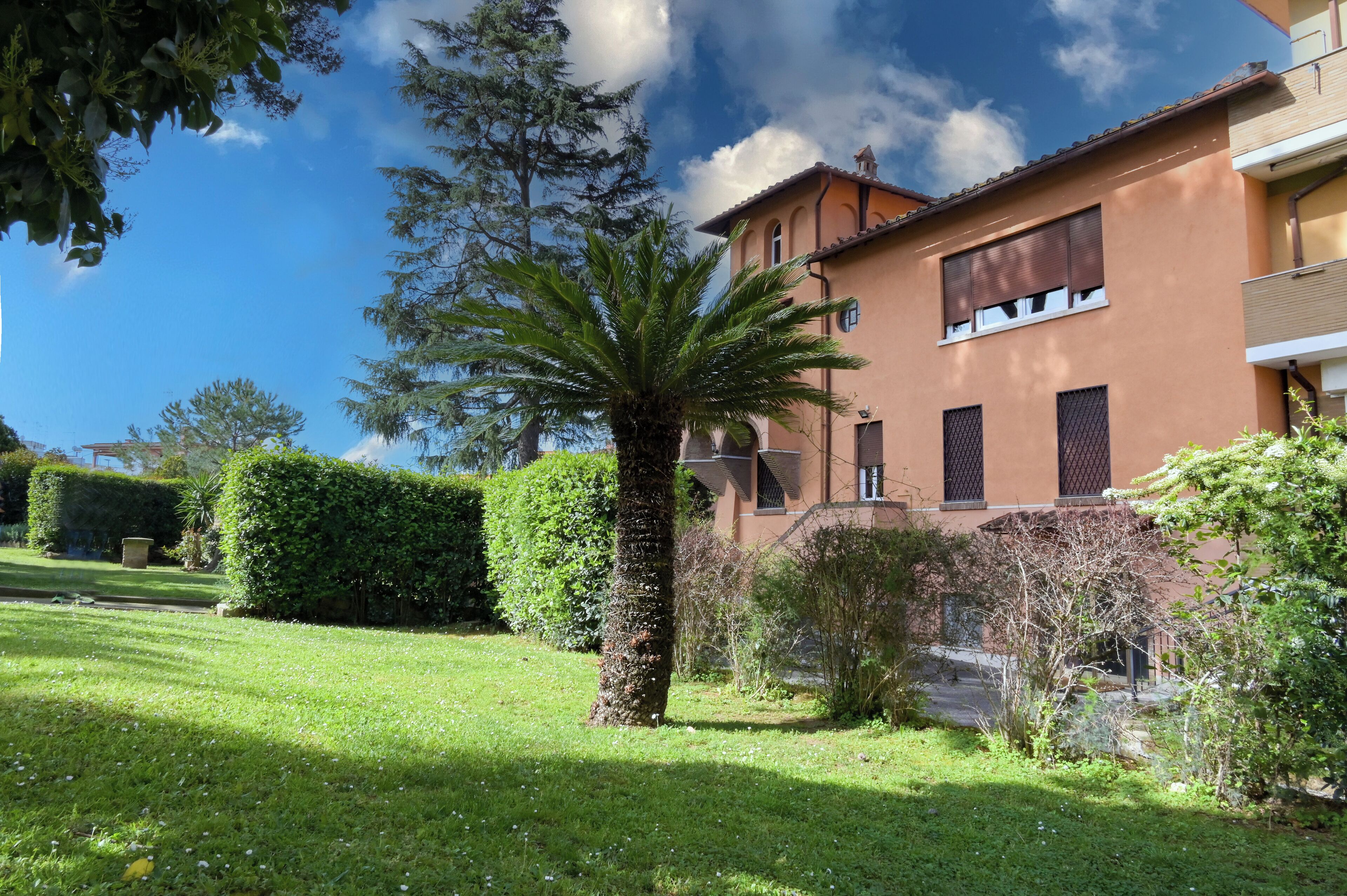 A building with a brown facade, multiple windows, and a well-maintained garden with a palm tree and hedges.