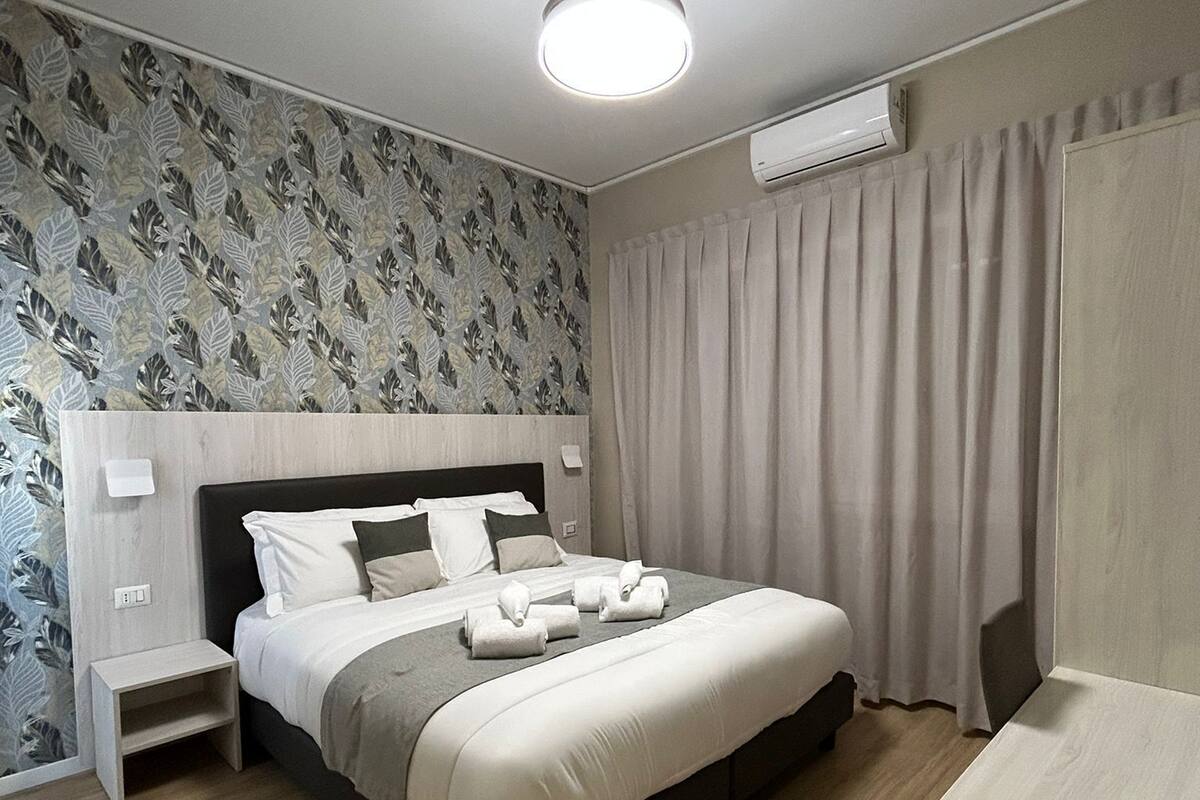 A modern hotel room with a neatly made bed, a bedside table, a small nightstand, and an air conditioning unit on the wall.