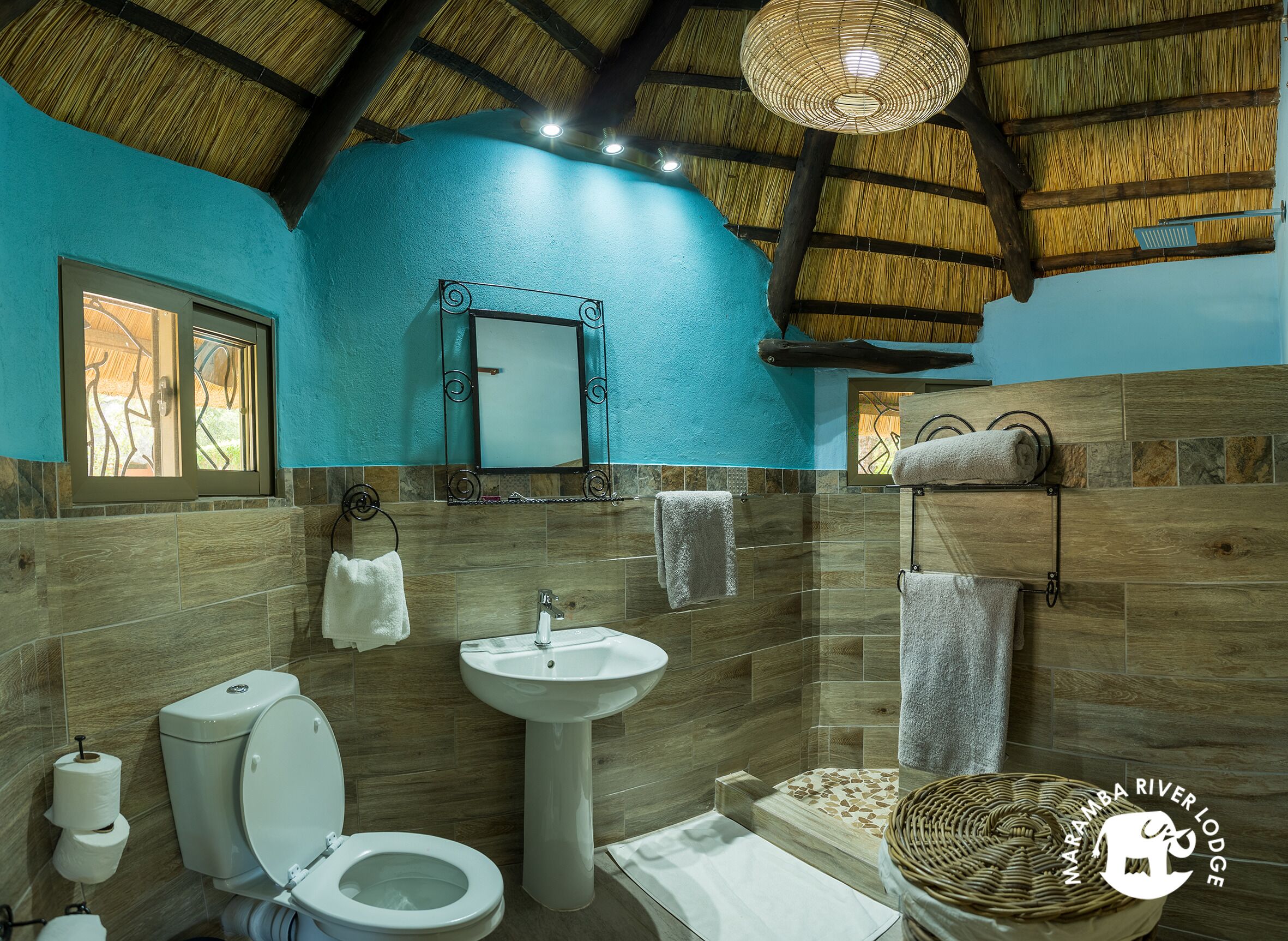 Un baño con inodoro, lavamanos y ducha. Cuenta con techo de paja, un espejo y toallas colgadas en la pared.