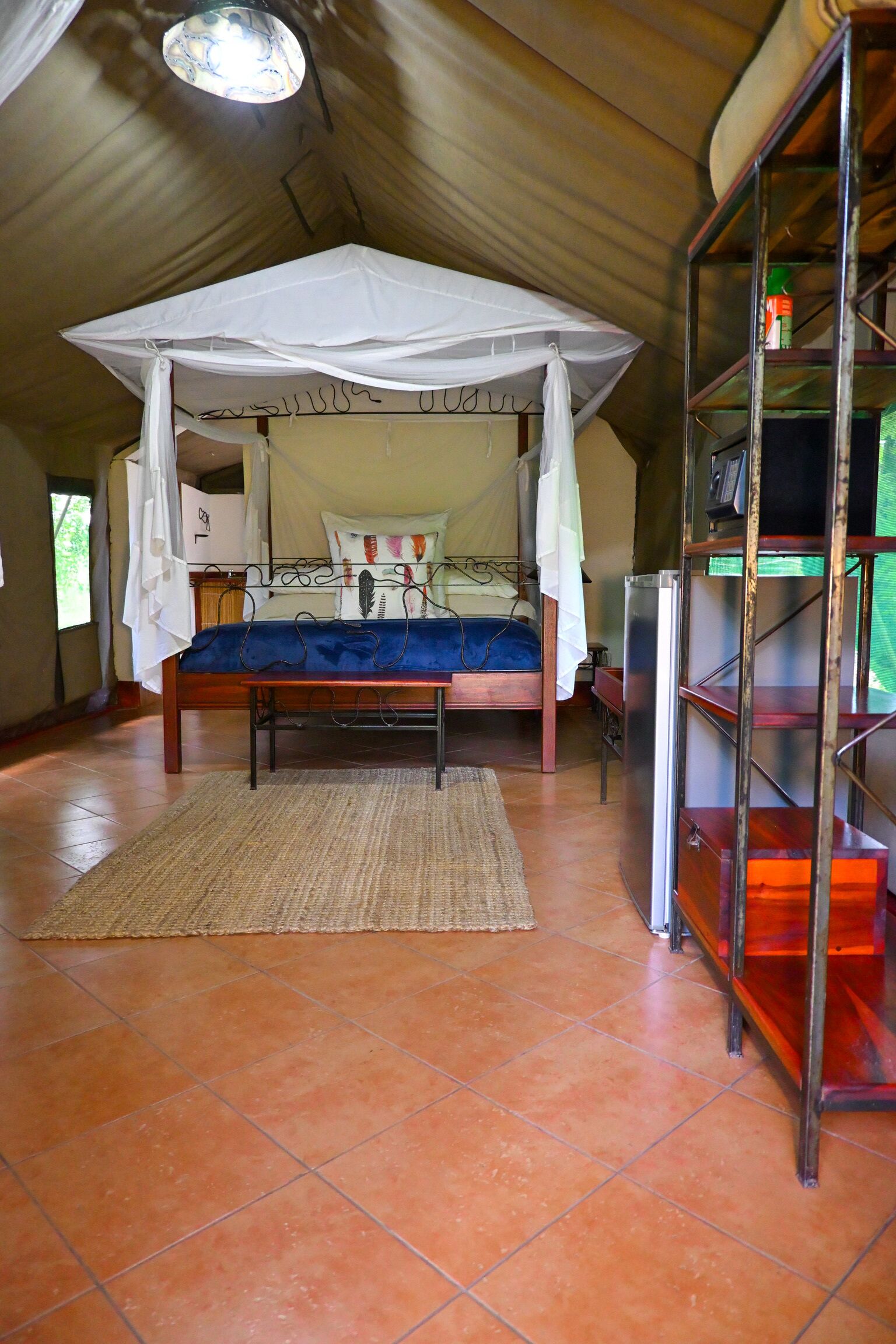 Ein Zimmer mit einem Bett unter einem Baldachin, einer Holzbank, einem Metallregal und einem Fliesenboden.