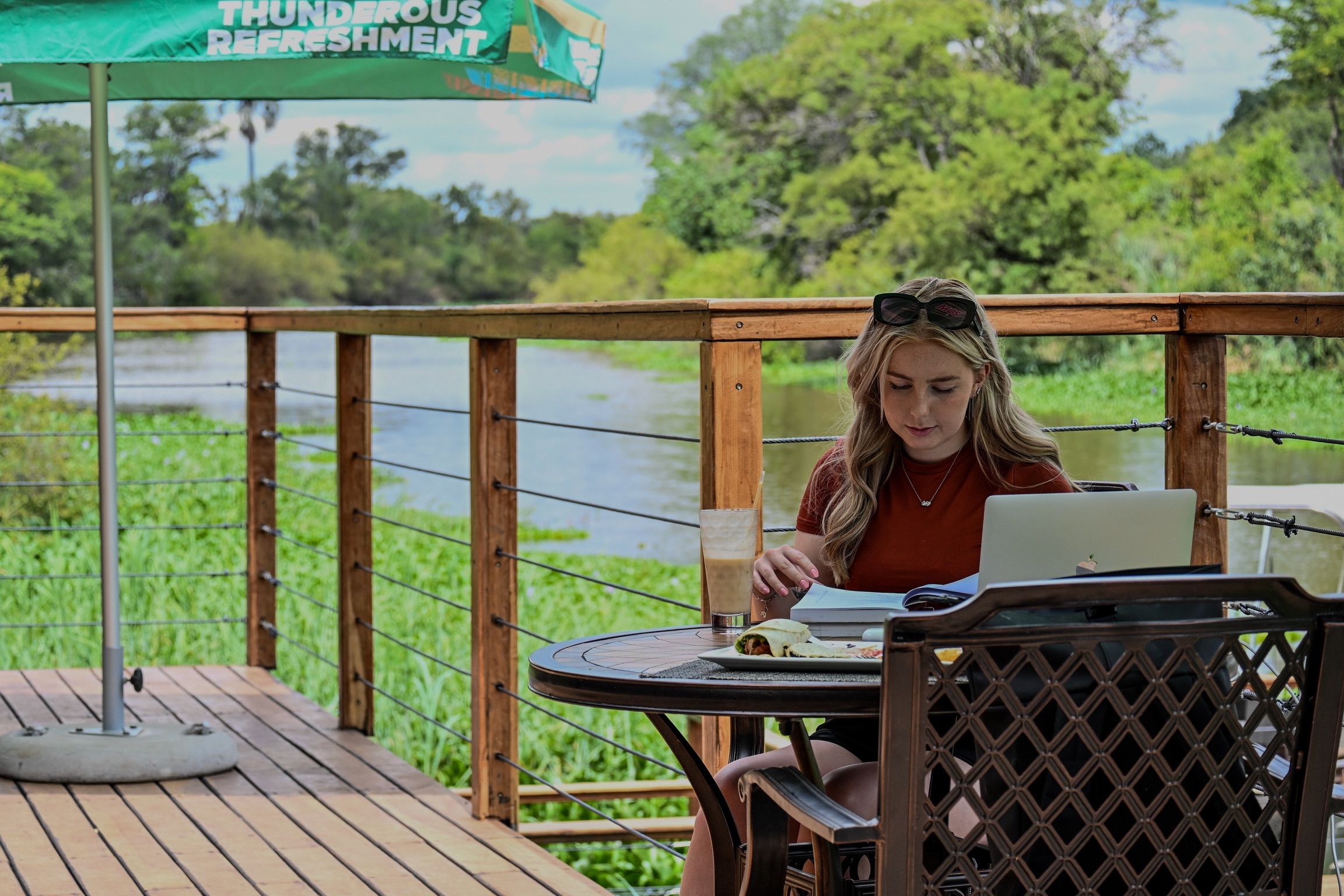 Une personne est assise à une table, devant un ordinateur portable, avec une boisson et une assiette de nourriture, sur une terrasse en bois offrant une vue sur une rivière et la verdure environnante.
