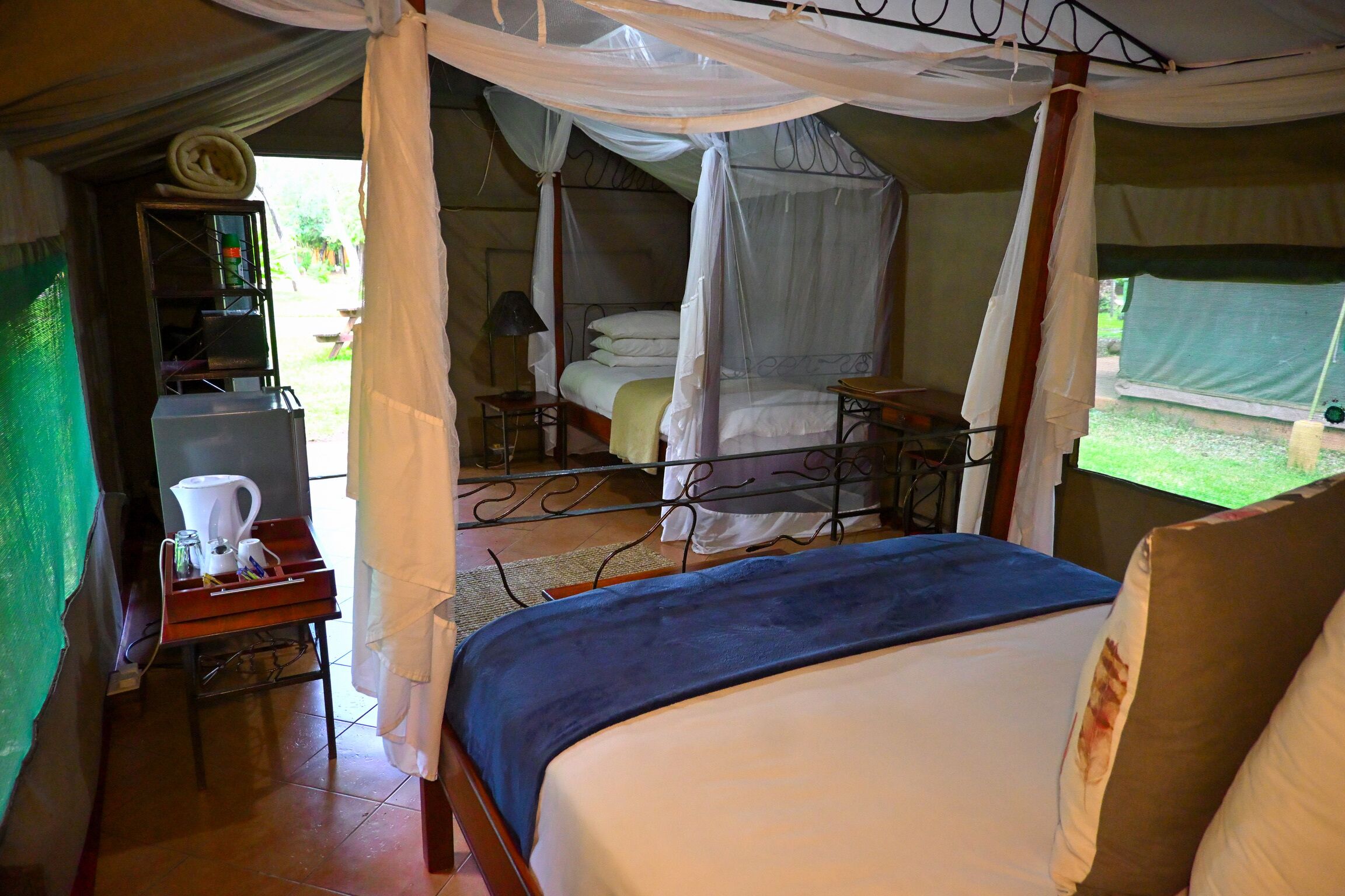 Alojamiento tipo tienda con una cama, una mesa con hervidor, y un estante pequeño con libros y una lámpara.