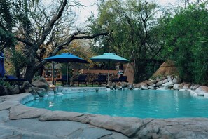 A pool area with lounge chairs and umbrellas, surrounded by rocks and trees. - Maramba River Lodge (Livingstone)