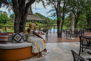 A person seated on a sofa with a view of a lake and trees. - Maramba River Lodge (Livingstone)