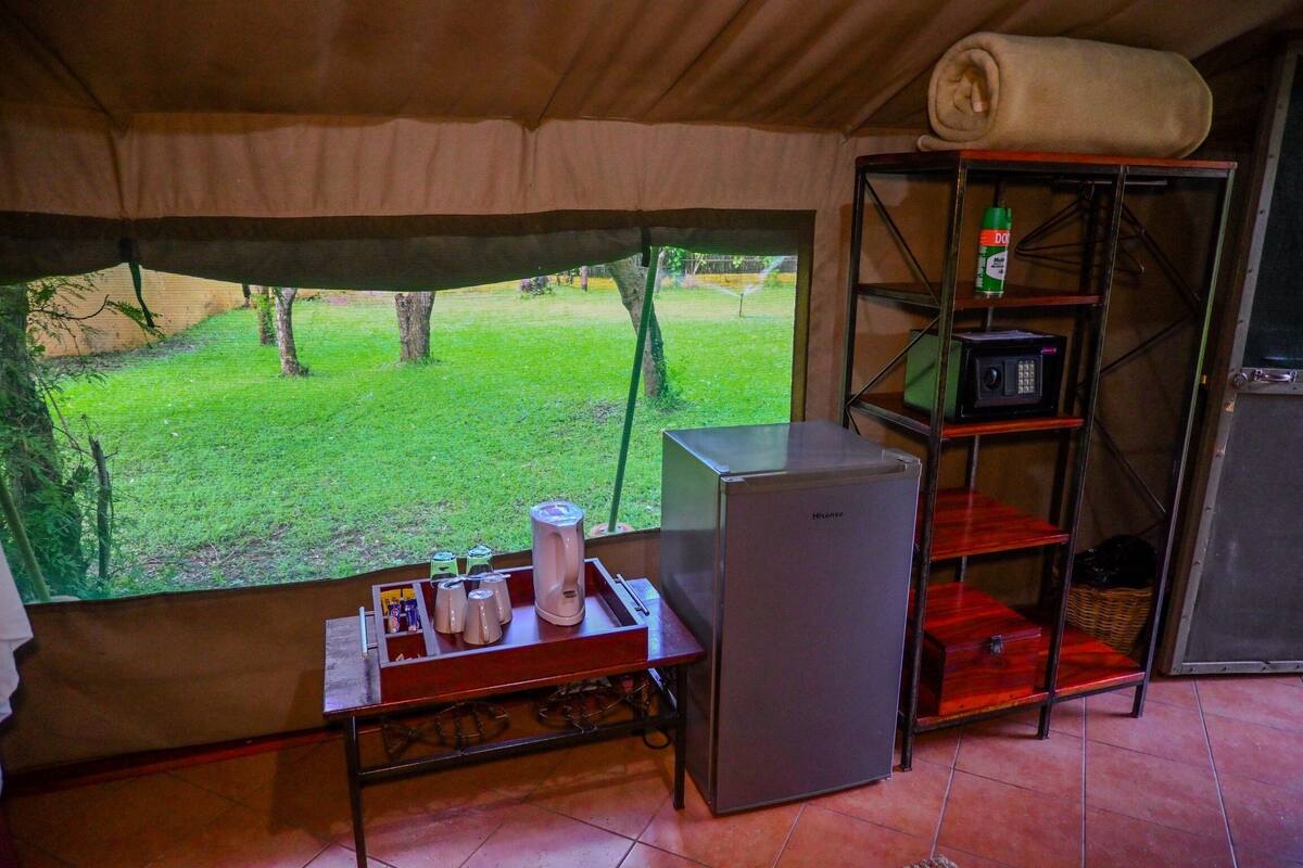 A compact kitchen with a refrigerator, a microwave, and a shelf with a bottle. A window reveals a grassy yard.