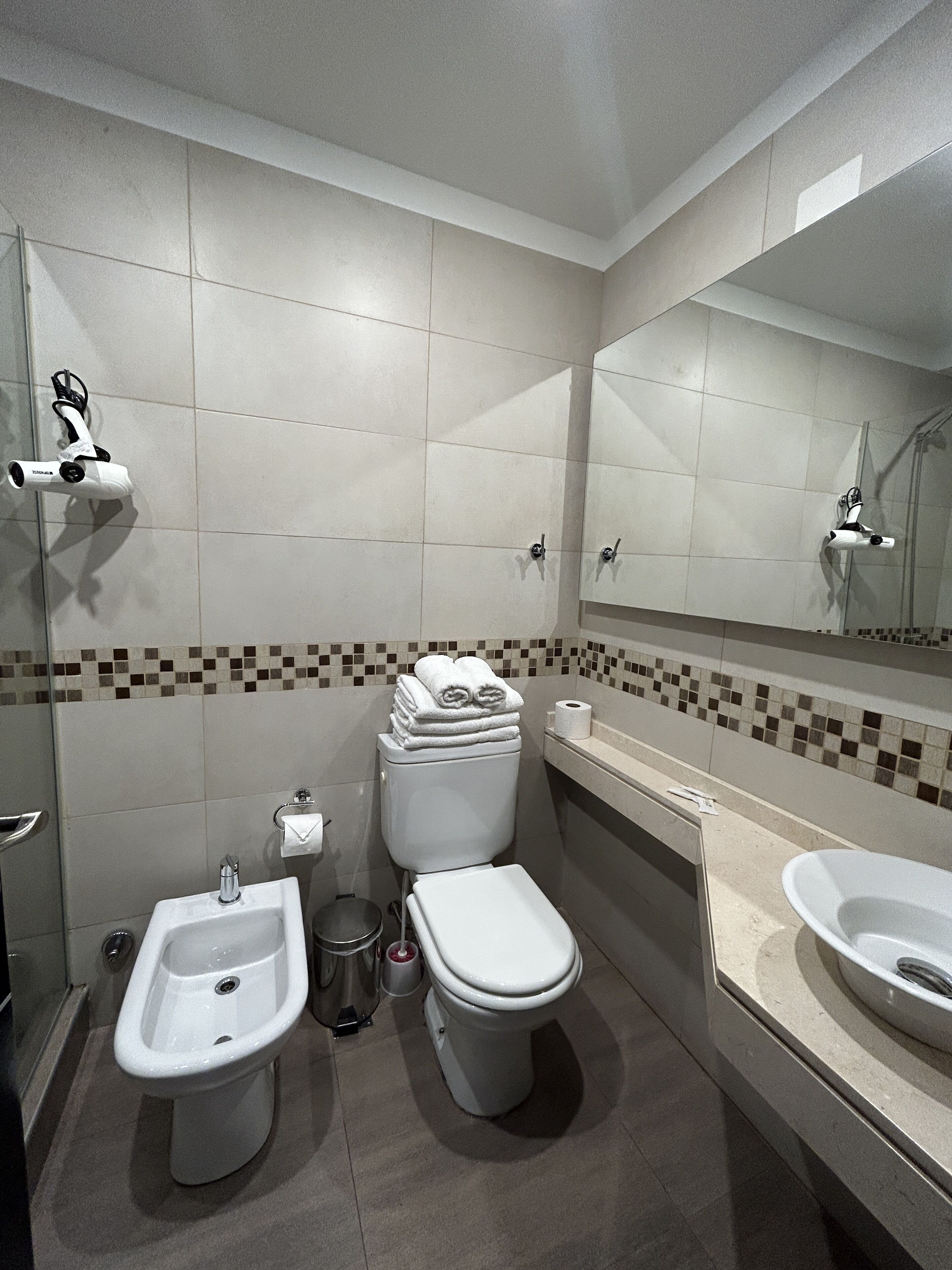 Un baño moderno con inodoro, bidé y lavamanos, paredes con azulejos y un espejo.