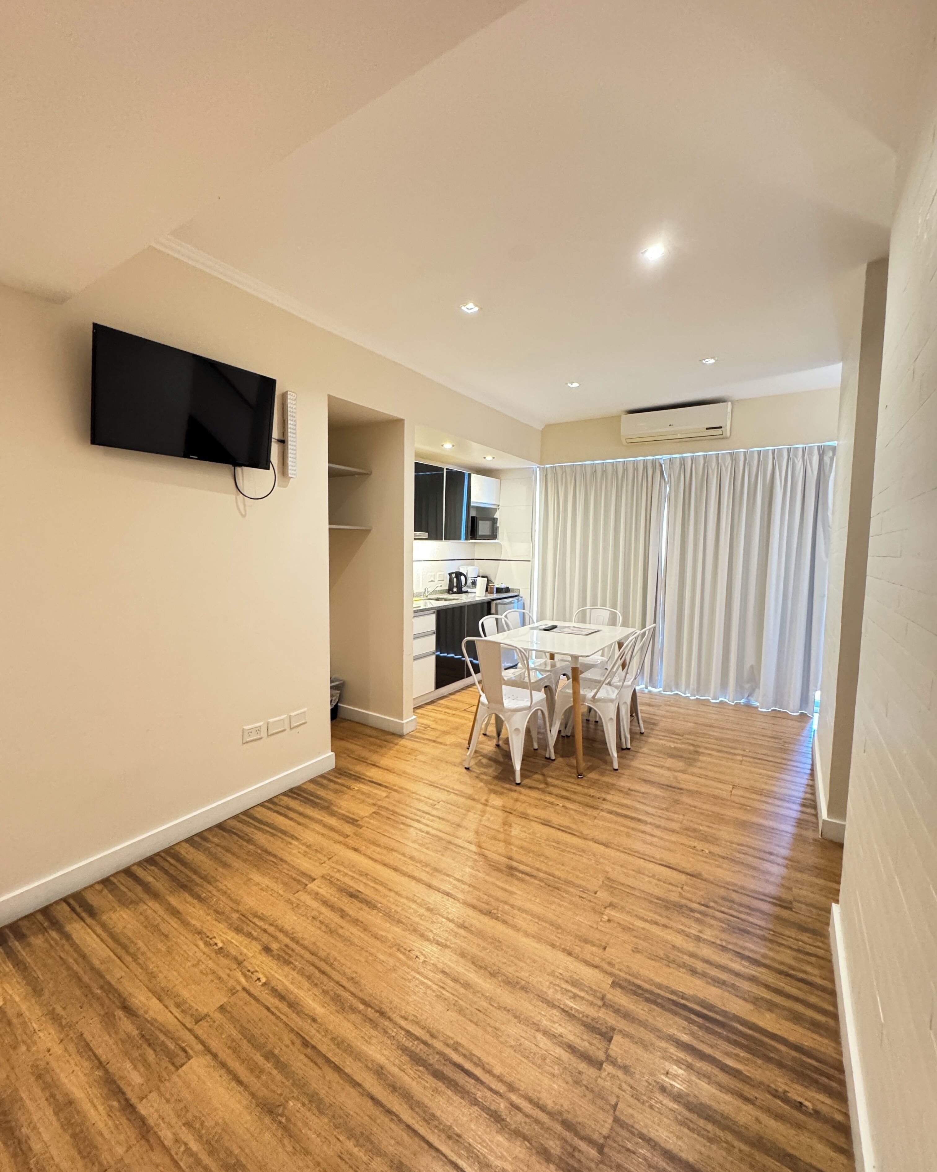 Een moderne woonkamer met een flatscreen televisie aan de muur, een eethoek met een witte tafel en stoelen, en een keuken met een magnetron en kasten.