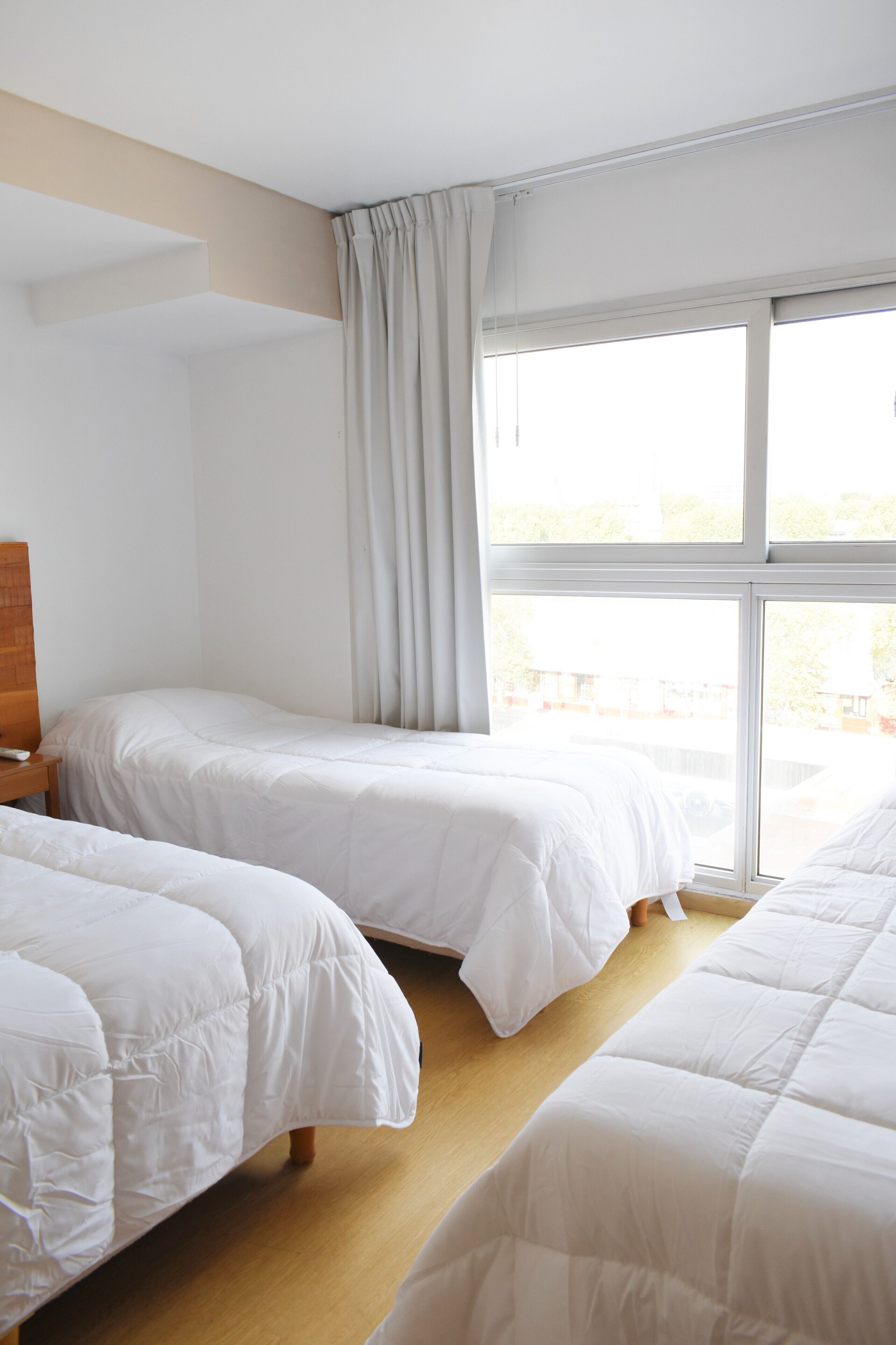 Quarto de hotel com duas camas de solteiro, uma mesinha de cabeceira de madeira, uma janela grande com cortinas e vista para o exterior.