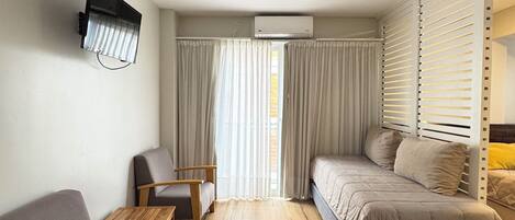 A compact hotel room with a bed, a sofa, a small table, and a chair. There is a window with curtains, an air conditioning unit, and a wall-mounted TV.