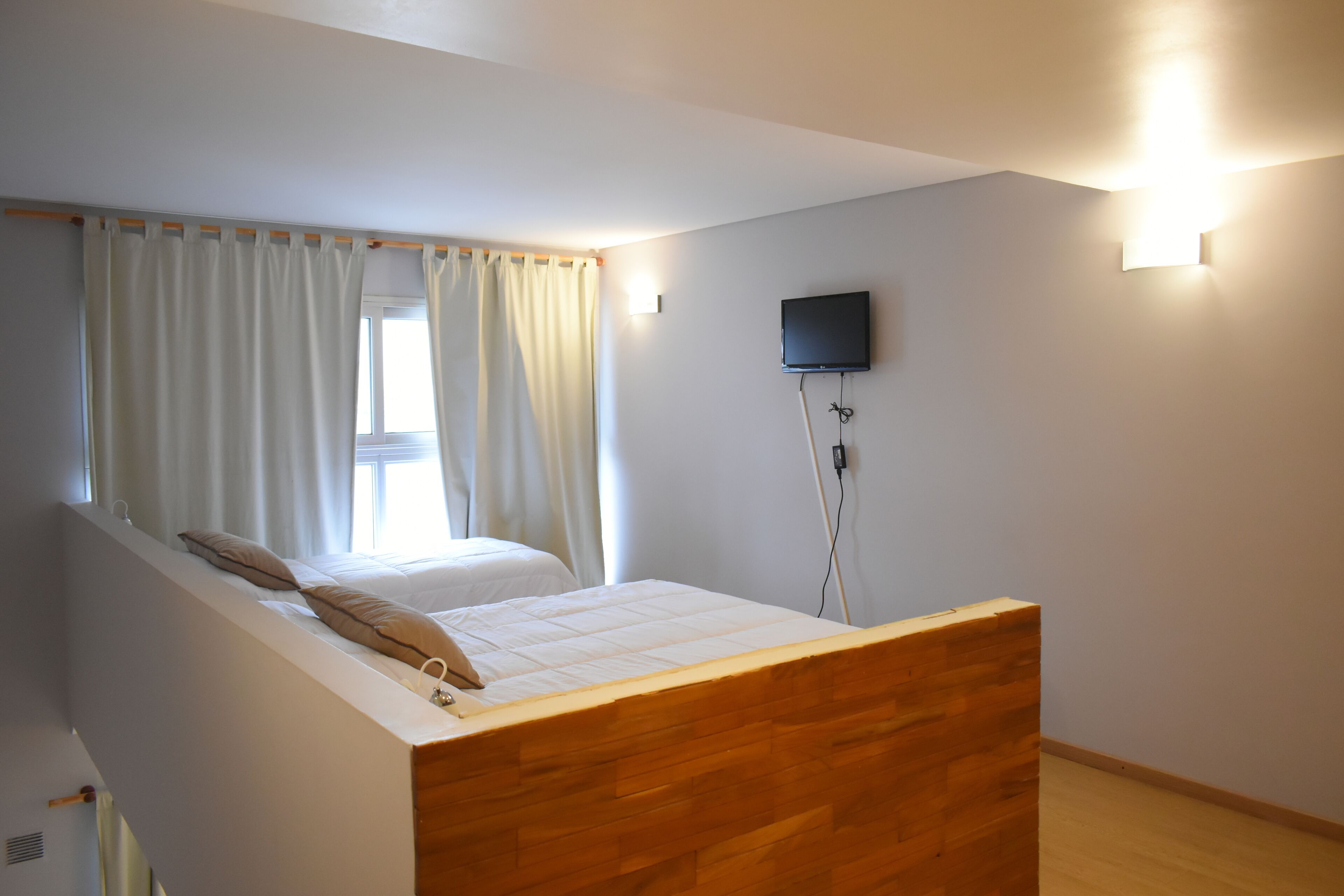 A bedroom with a wooden headboard, a bed with white bedding, a window with curtains, a wall-mounted TV, and a light fixture.