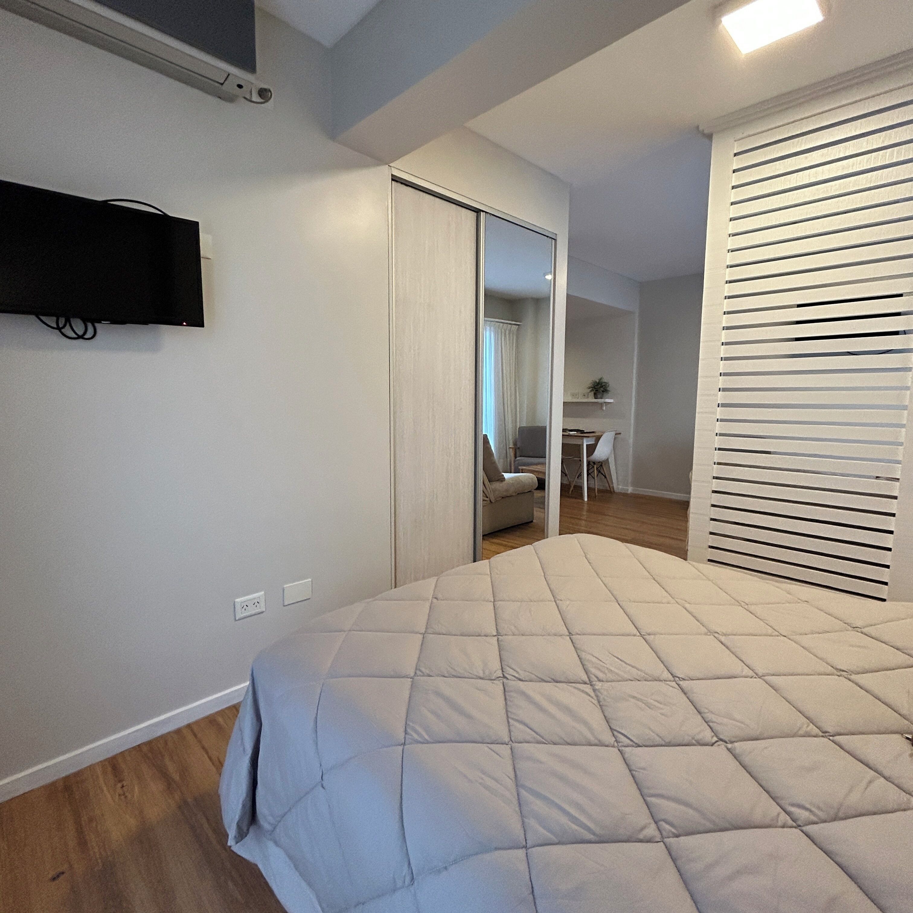 A bedroom with a bed, a TV mounted on the wall, a desk, and a chair in the adjacent room.