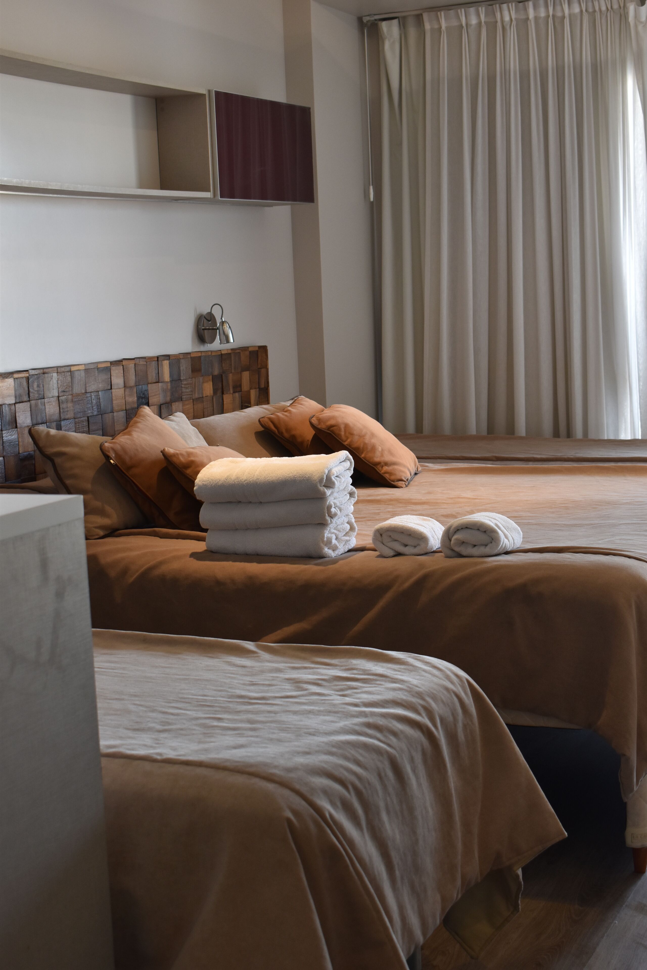 A hotel room with two beds, beige bedding, a wooden headboard, a bedside table, and a window with curtains.