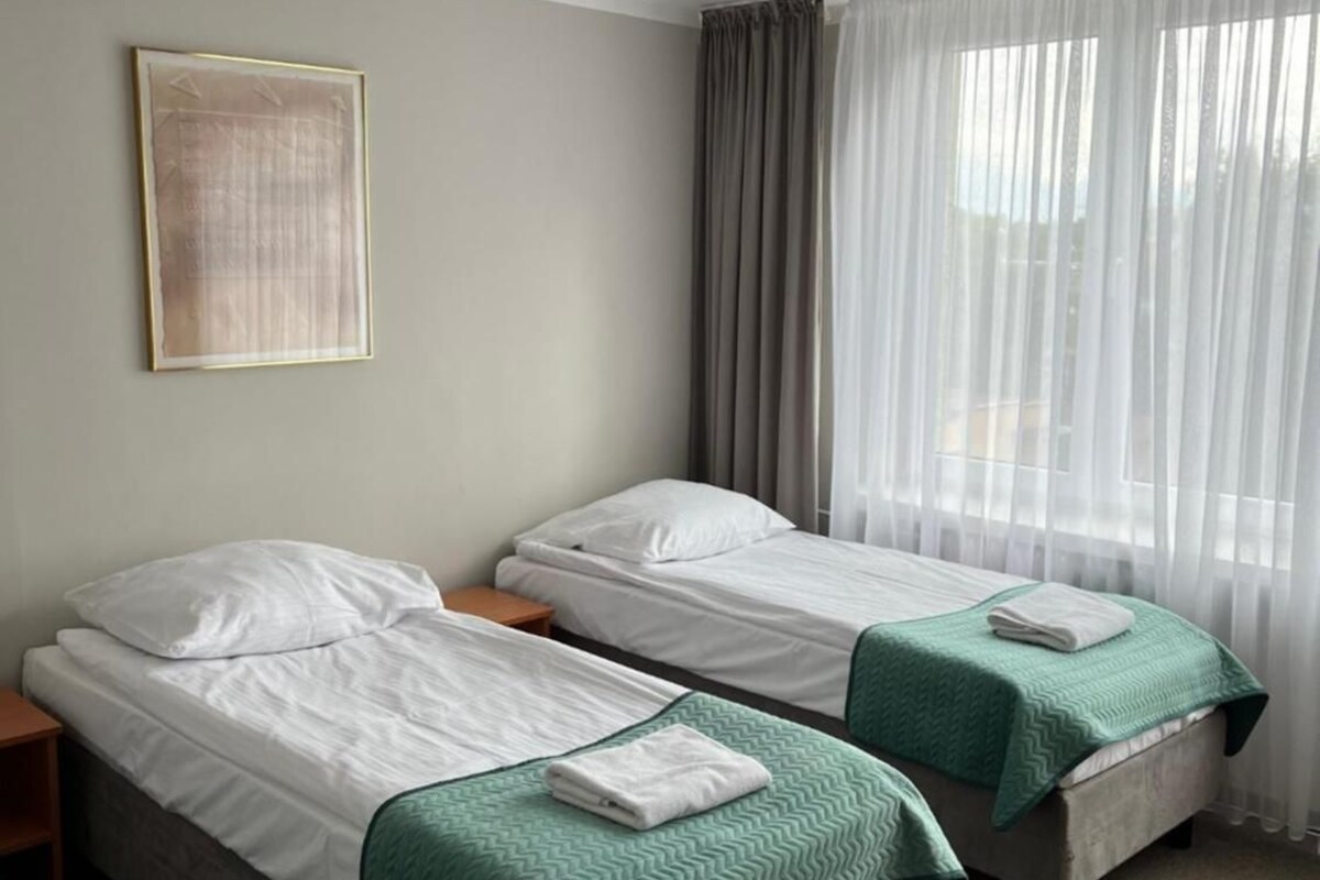 A hotel room with two beds, a ceiling light, a window with curtains, and a framed picture on the wall.