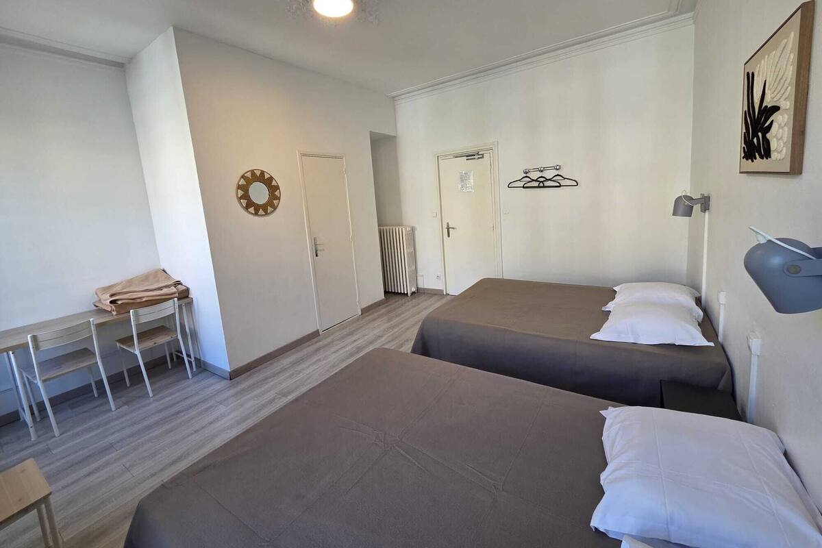 A hotel room with two beds, a desk, a chair, a radiator, and a wall-mounted lamp.