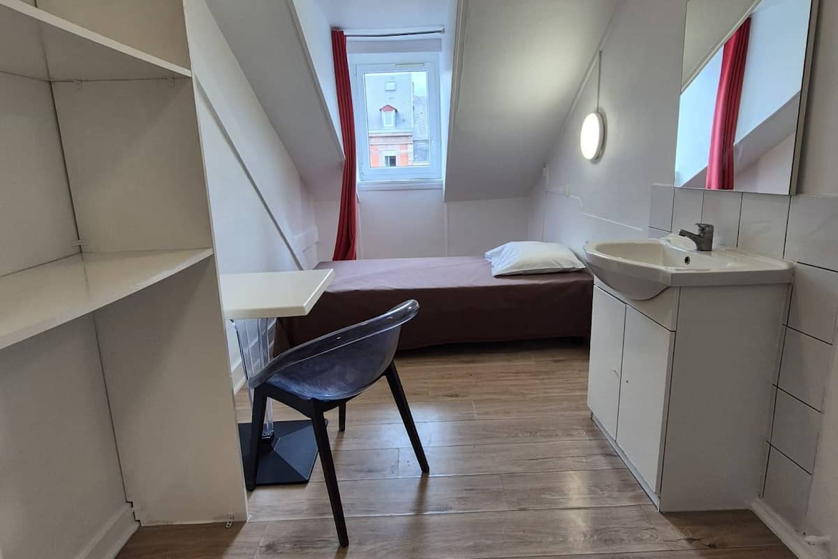 A compact, modern bedroom with a sloped ceiling, a skylight, a single bed, a desk, and a chair.