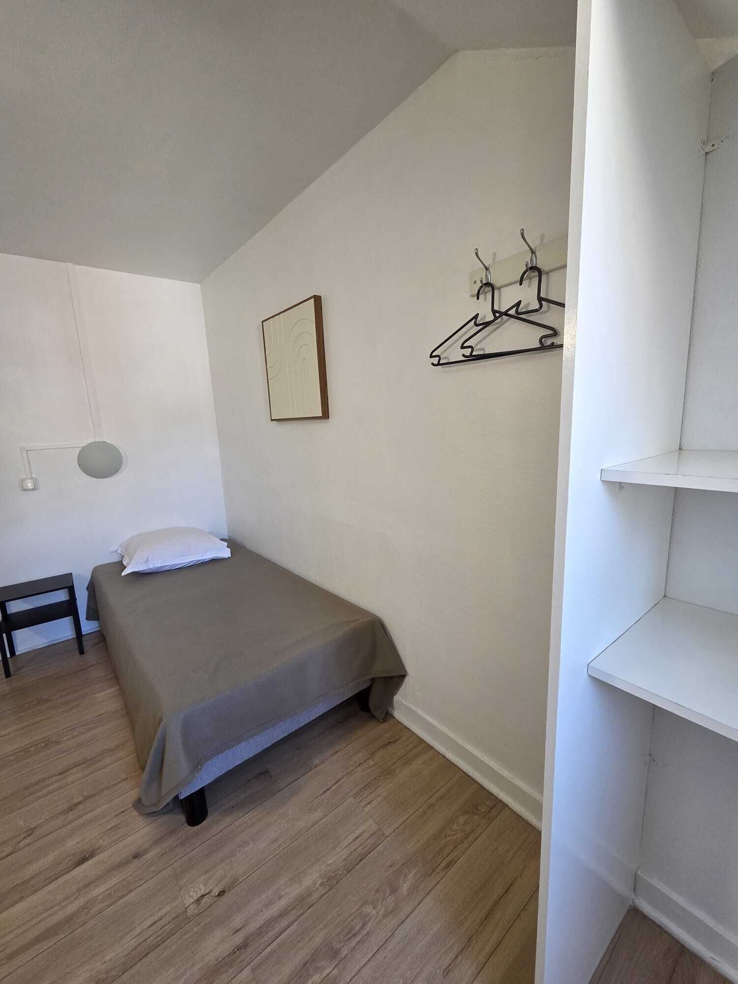 A bedroom with a bed, a bedside table, a hanging light fixture, a wall-mounted coat rack, and a shelf.