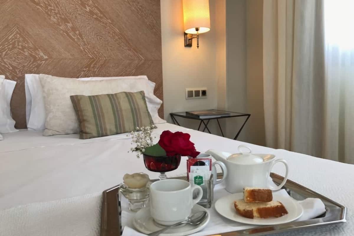Ein ordentlich bezogenes Bett mit Kissen, ein Nachttisch mit Lampe und ein Tablett mit Frühstücksgebäck, darunter eine Tasse, ein Teekessel und ein Stück Kuchen.