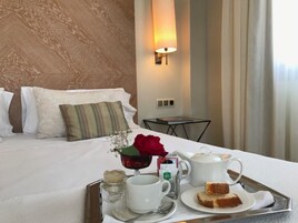 Una cama bien hecha con almohadas, una mesita de noche con una lámpara y una bandeja con productos para el desayuno, incluyendo una taza, una tetera y una porción de pastel.