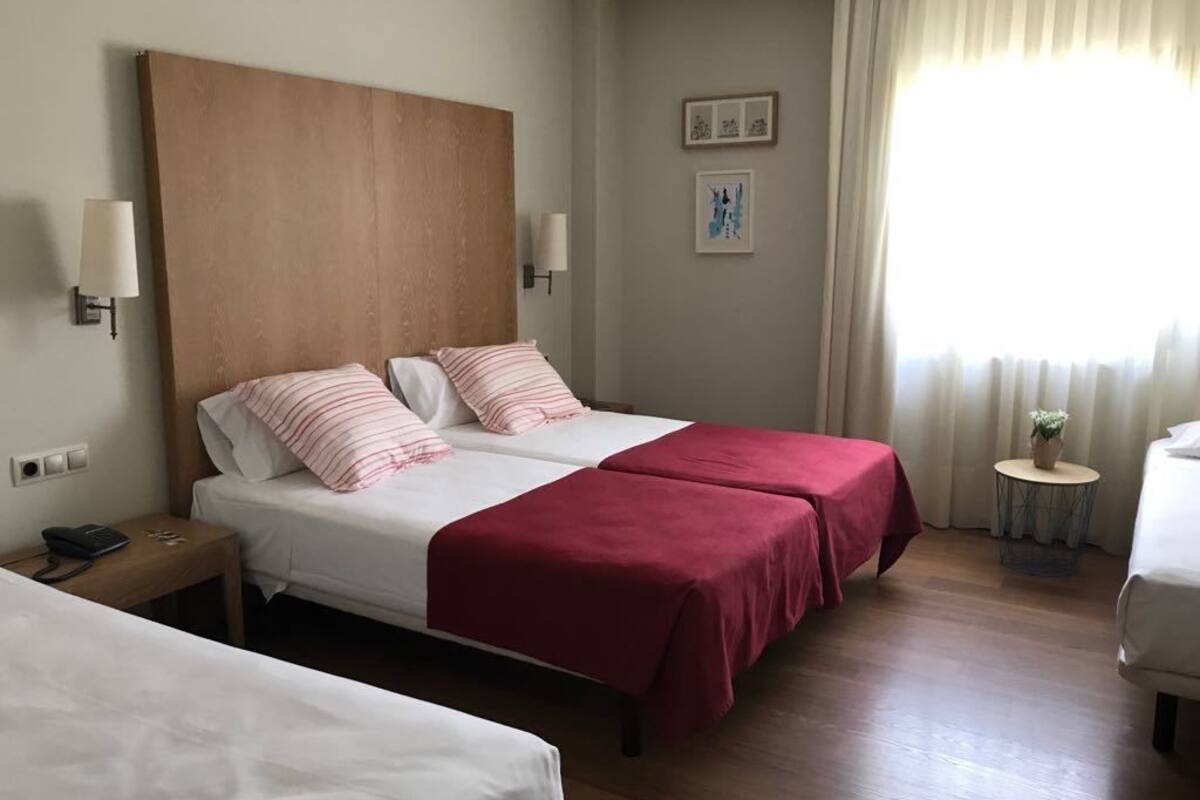 Ein Hotelzimmer mit zwei Betten, einem Holzkopfstück, einem kleinen Nachttisch, einer Lampe und einem Fenster mit Vorhängen.