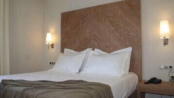 A hotel room with a large bed, two bedside lamps, a wooden headboard, and a nightstand with a phone and a bag.