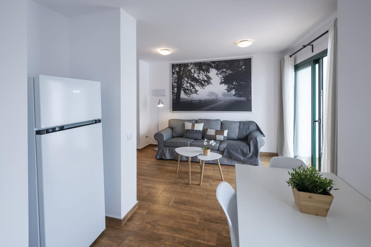 Un salon moderne avec un canapé gris, une table à manger blanche et un coin cuisine doté d’un réfrigérateur blanc.