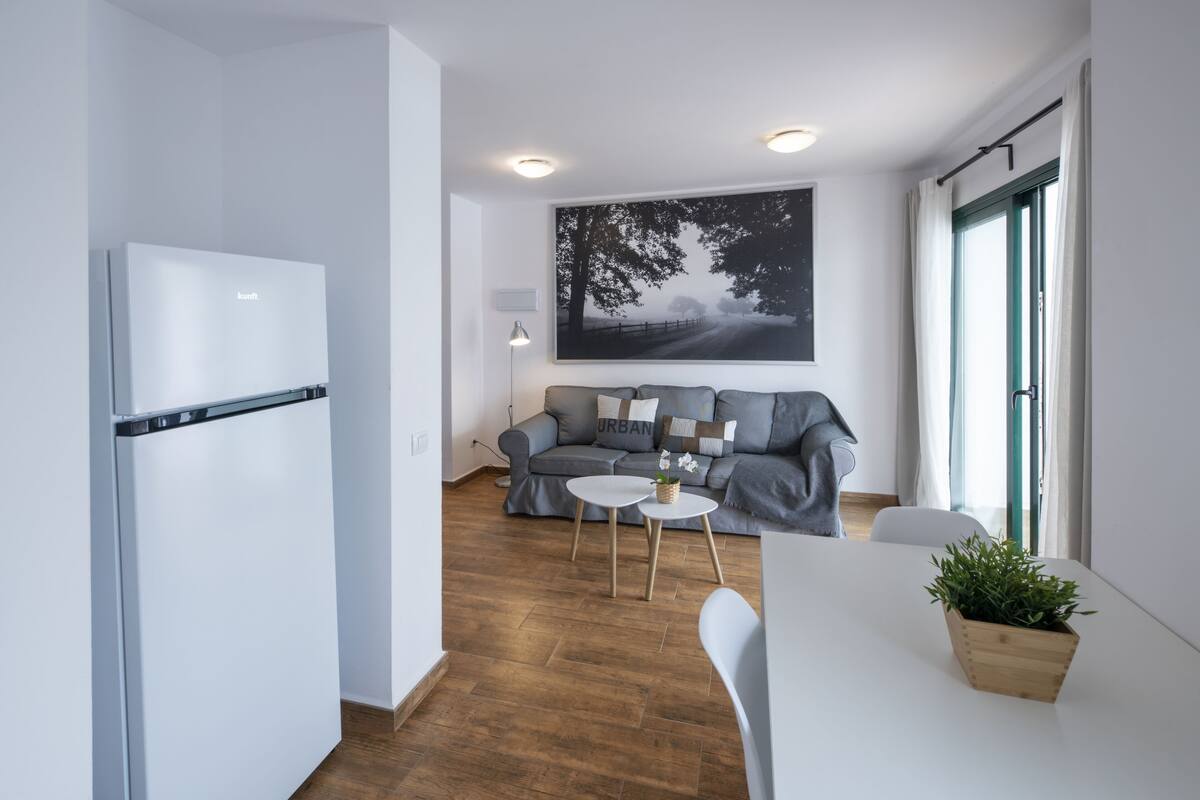 Ein modernes Wohnzimmer mit einem grauen Sofa, einem weißen Esstisch und einer Küchenzeile mit einem weißen Kühlschrank.
