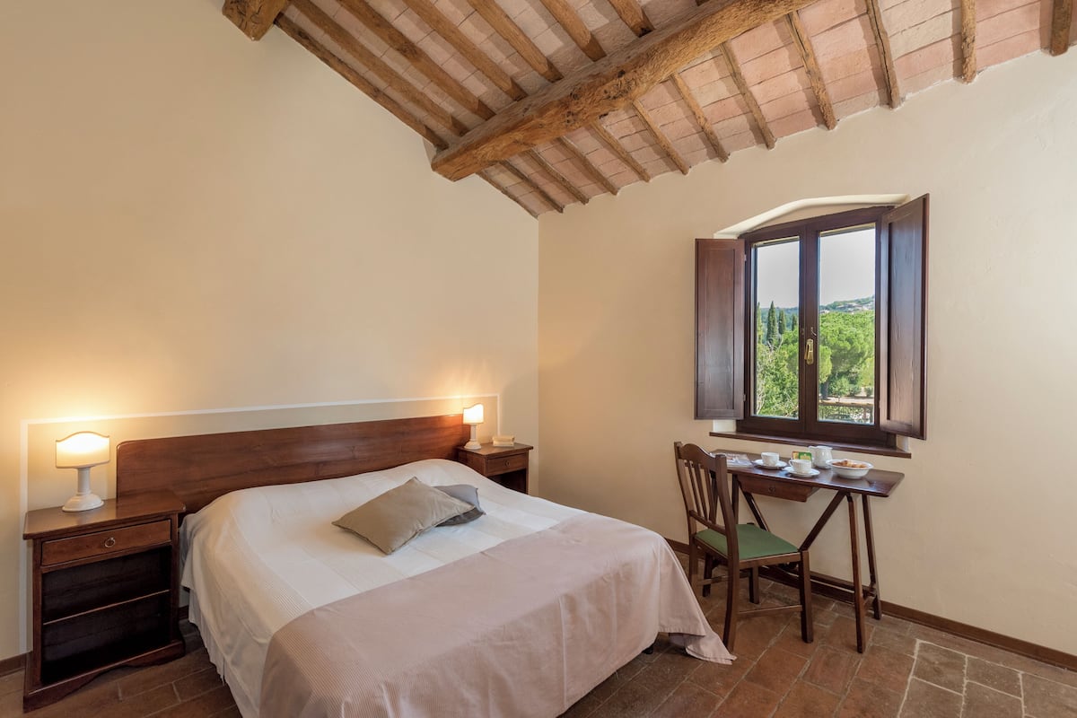 A bedroom with a wooden headboard, a bedside table with a lamp, a desk with a chair, and a window with a view of greenery.