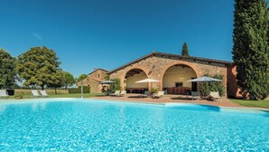 A swimming pool with lounge chairs and umbrellas, a stone building with arches, and a large tree in the background. - Country House Le Case Coloniche (Deruta)