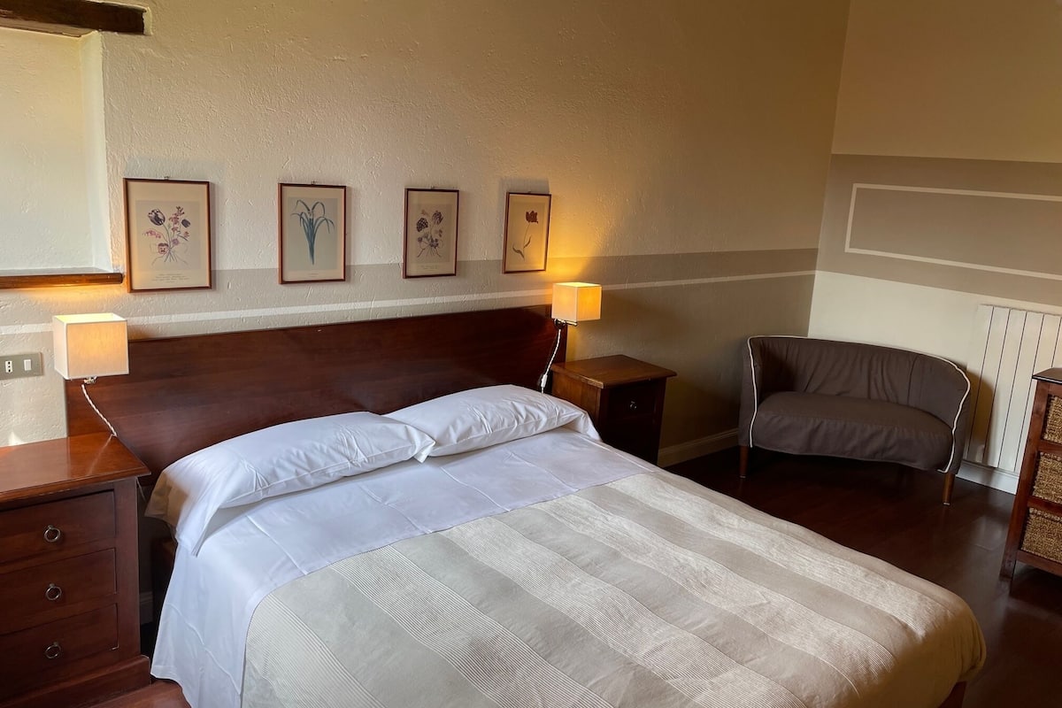 A neatly made bed with a striped bedspread, a wooden headboard, and a nightstand with a lamp. There are framed pictures on the wall and a chair in the corner.