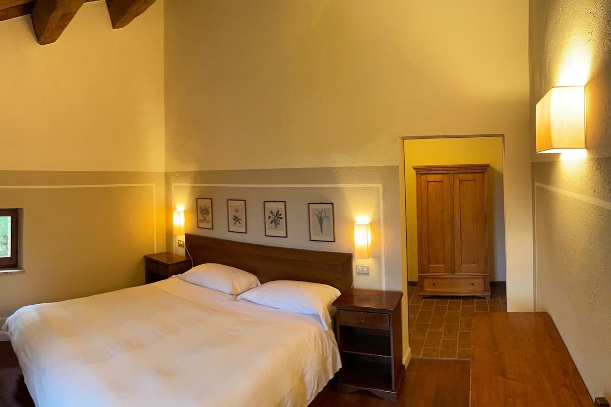 A bedroom with a large bed, wooden headboard, bedside tables, wall-mounted lamps, framed pictures, and a wooden wardrobe.
