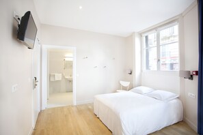 A bedroom with a bed, a TV mounted on the wall, a window with a view of buildings, and a door leading to a bathroom. - Nadaud Hôtel (Paris)