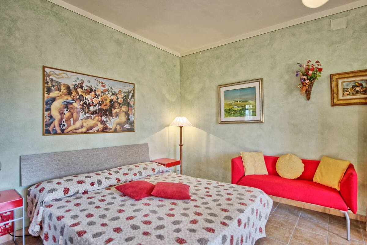 A bedroom with a bed, a red sofa, a nightstand, a lamp, and two paintings on the wall.