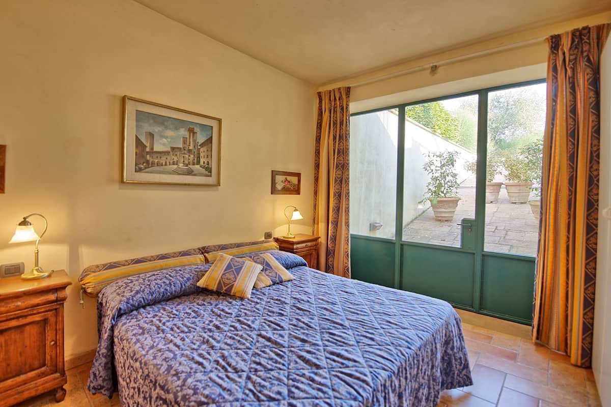 A bedroom with a large bed, two bedside tables, a nightstand, a window with curtains, and a painting on the wall.