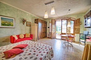 A room with a bed, a red sofa, a dining table with chairs, wooden cabinets, and a view through an open door.