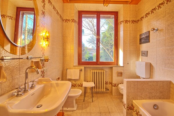 Un baño con lavamanos, inodoro y bañera, una ventana con vista a árboles, y un espejo.