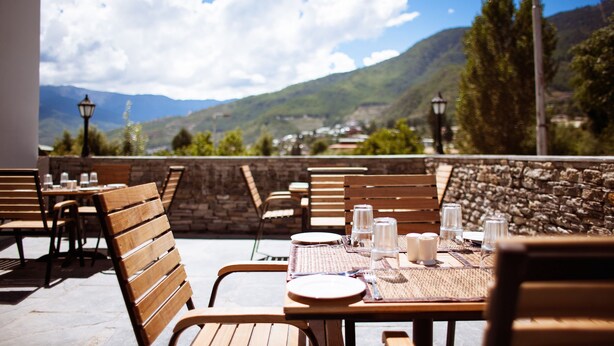 An outdoor dining area with wooden tables and chairs, set against a backdrop of mountains and a clear sky. - Hotel Osel (Thimphu)
