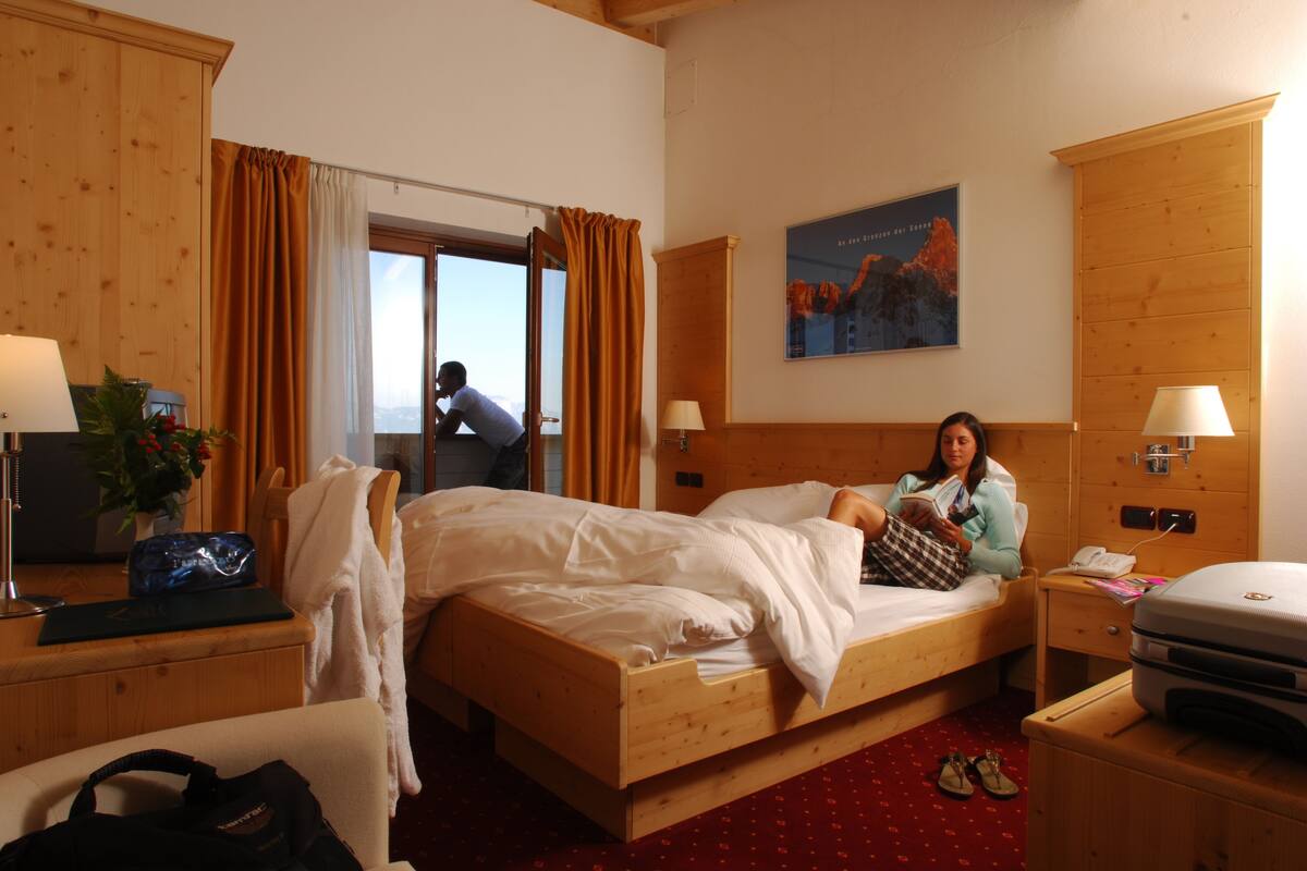Une chambre d’hôtel avec un lit en bois, un bureau avec une lampe, une chaise, une valise et une fenêtre avec des rideaux.