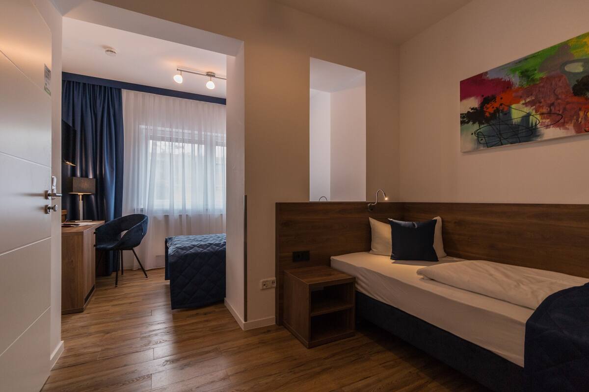 A modern hotel room with a wooden floor, a bed with white linens, a desk with a chair, and a painting on the wall.