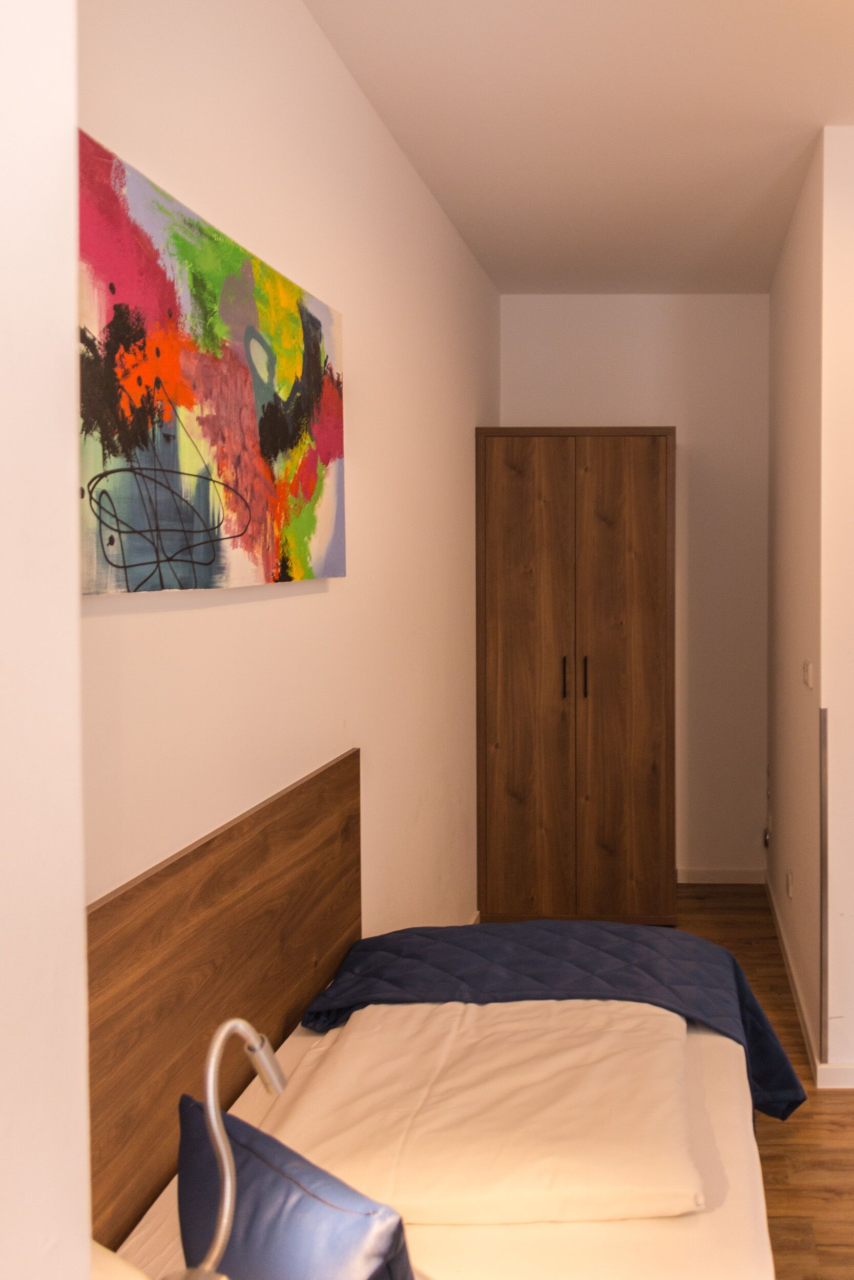 A bedroom with a bed, a wooden wardrobe, and a colorful abstract painting on the wall.