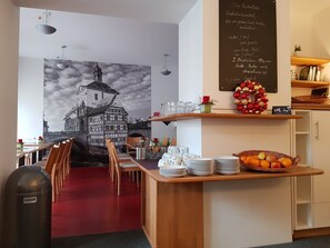 Interior de un restaurante con un mural de un pueblo europeo, una pizarra con el menú y una cesta de frutas en la barra.