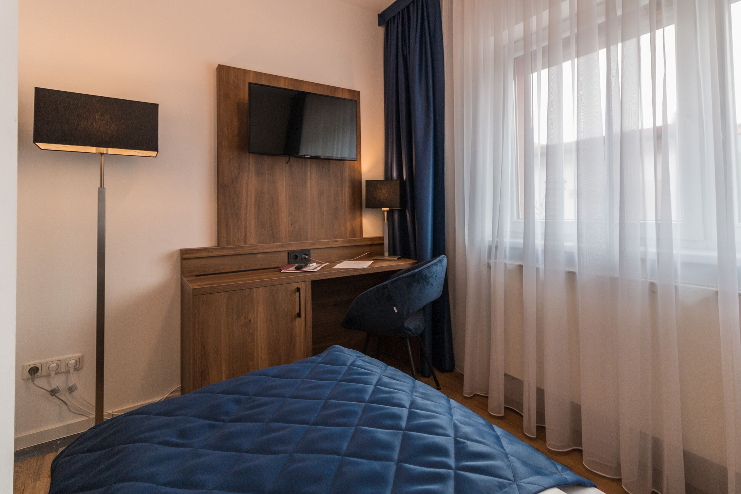 A hotel room with a bed, a desk, a chair, a TV on the wall, and sheer curtains.